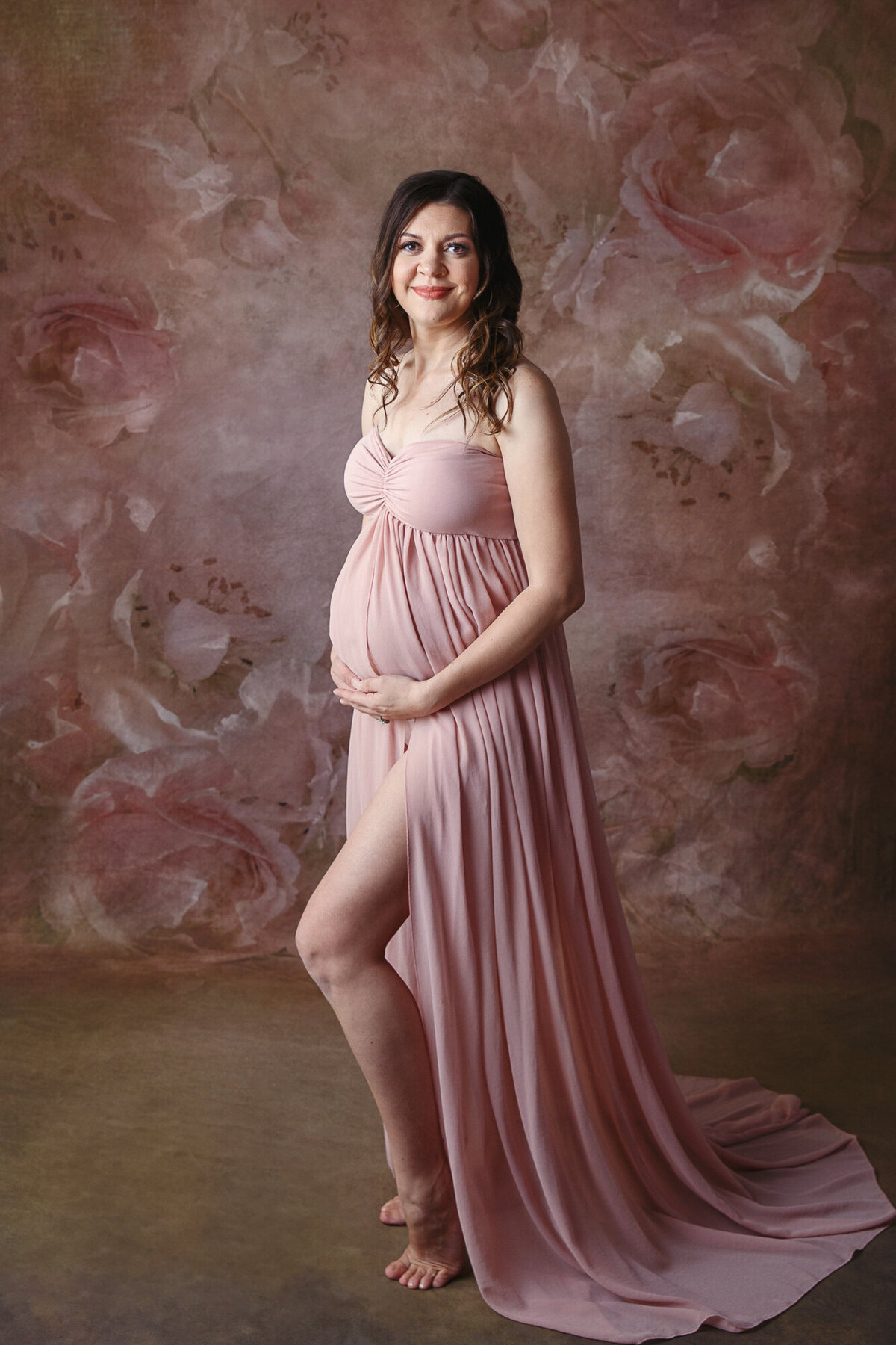 Pregnant woman standing with hands under her baby belly wearing a pink maternity gown on a floral pink and brown background