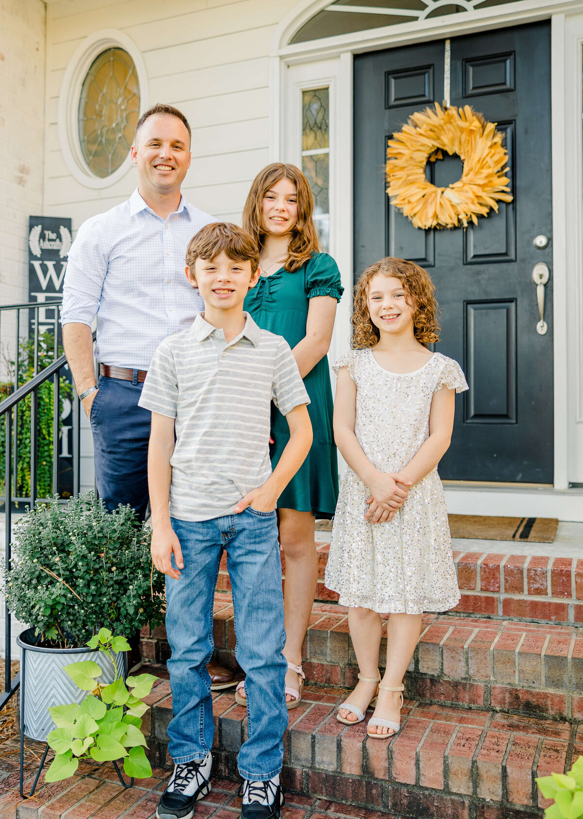 In front of a house with a black door adorned with a wreath, a family of four beams happily. Captured by a Fayetteville NC family photographer, the father stands on the left while two girls in dresses and a boy in a striped shirt stand before blooming plants on the steps.