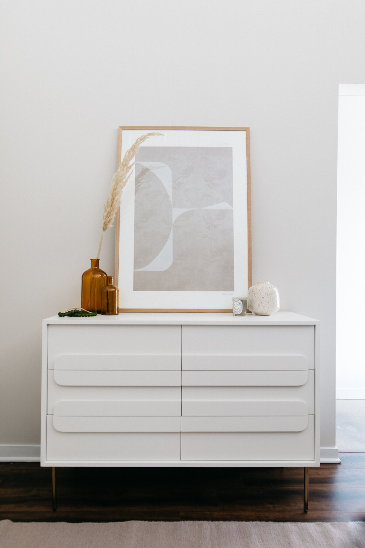 White double dresser with oversize drawer handles. Abstract grey beige wood framed artwork on top leaning against wall. Amber glass bottle vases styled with pampas grass and candle and ceramic vasein front