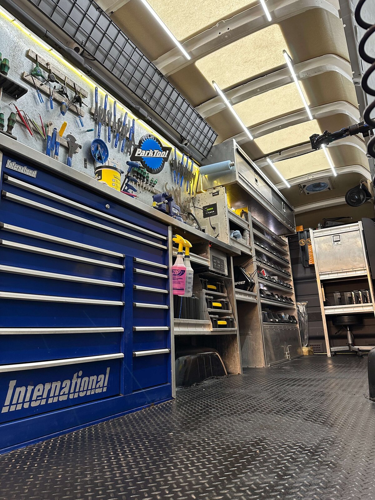 A well-organized workshop inside a van with toolboxes and various tools neatly arranged on shelves and racks. The interior features a patterned floor and a ceiling with striped lighting