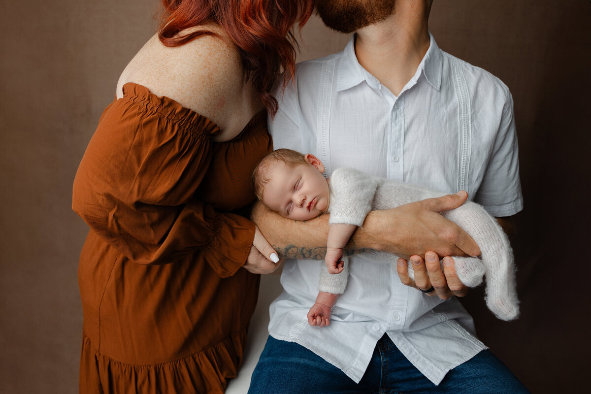 Newborn baby cradled in dad’s arms as mom leans in close during their studio session, styled in warm and neutral tones. Captured by Inland Empire newborn photographer Kaitlyn Dawn Photography.