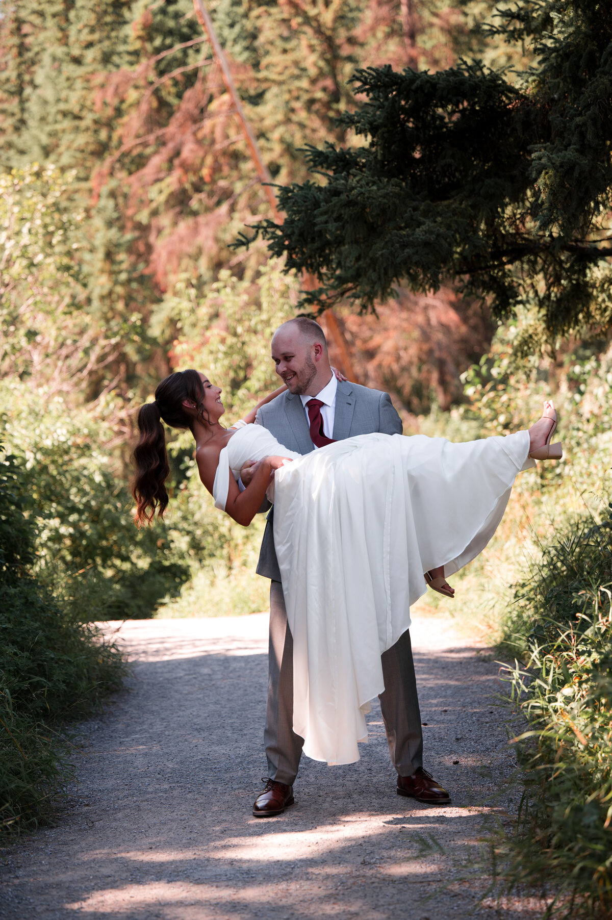 groom carries his bride down a sunny pathway in the forest