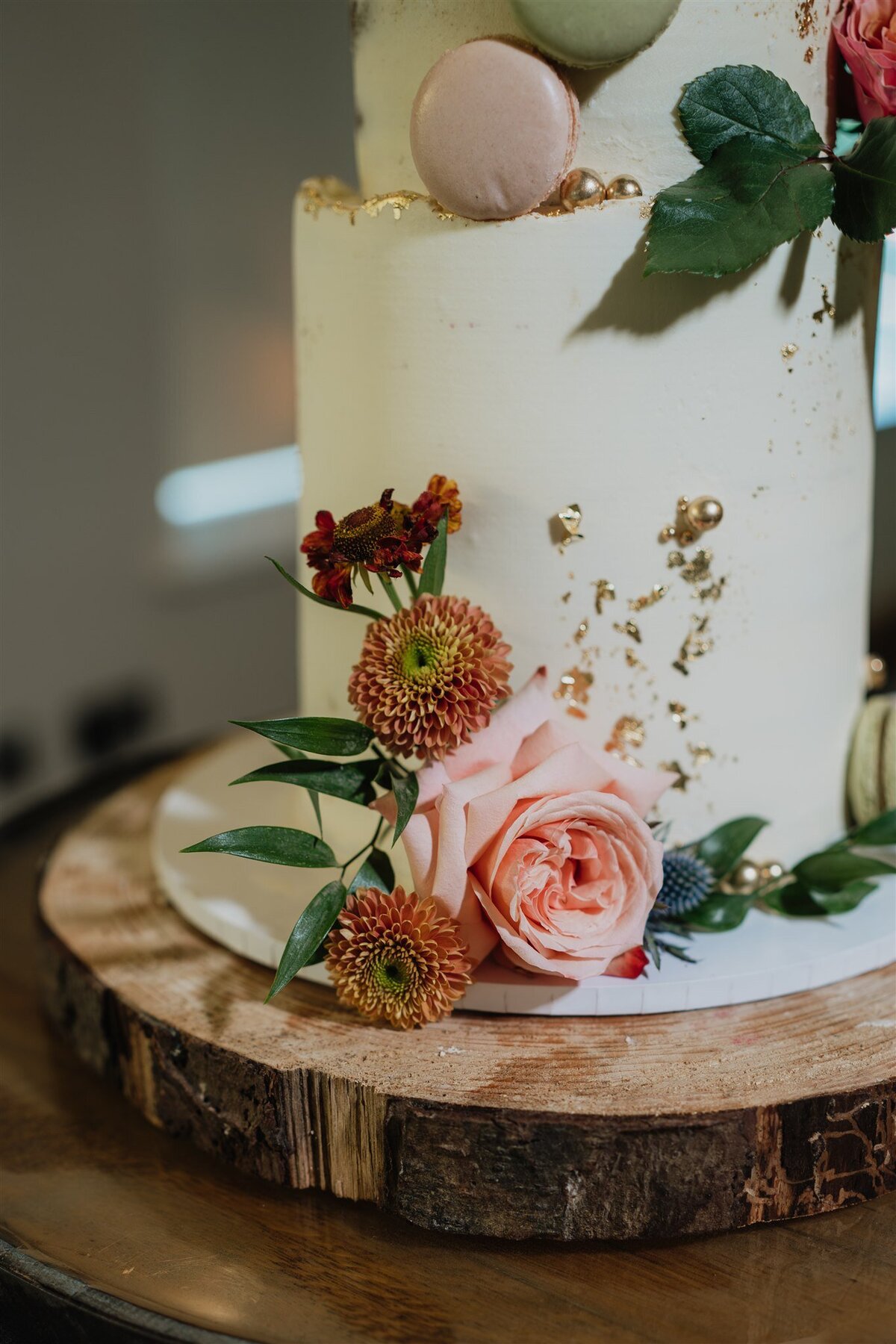 A three tier wedding cake stands on a rustic barrel. The cake is decorated with cream icing, roses, macarons, and foliage.