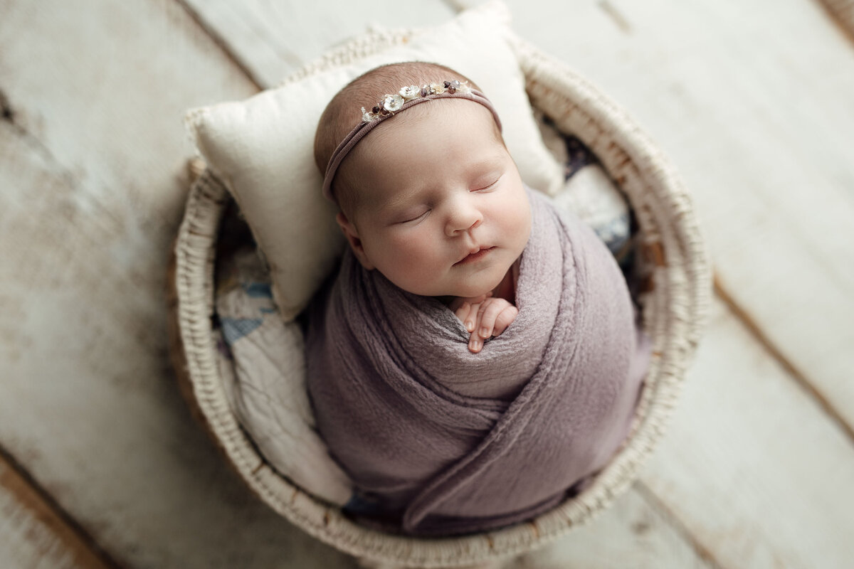 Baby sleeping in basket on top of a cream coloured painted hardwood floor.   Baby is wrapped in a pale lilac wrap with petite headband with tiny flowers.