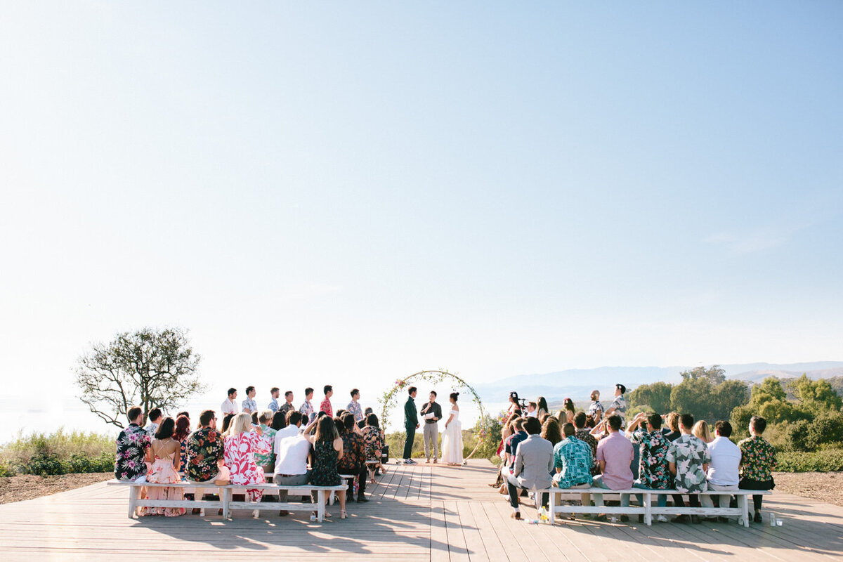 A wide shot of the entire wedding party and ceremony as the bride and groom stand in front of the gold arch and flowers at Dos Pueblos Orchid Farm. The guests wear casual tropical themed attire, by SoCal photographer My Sun and Stars Co.