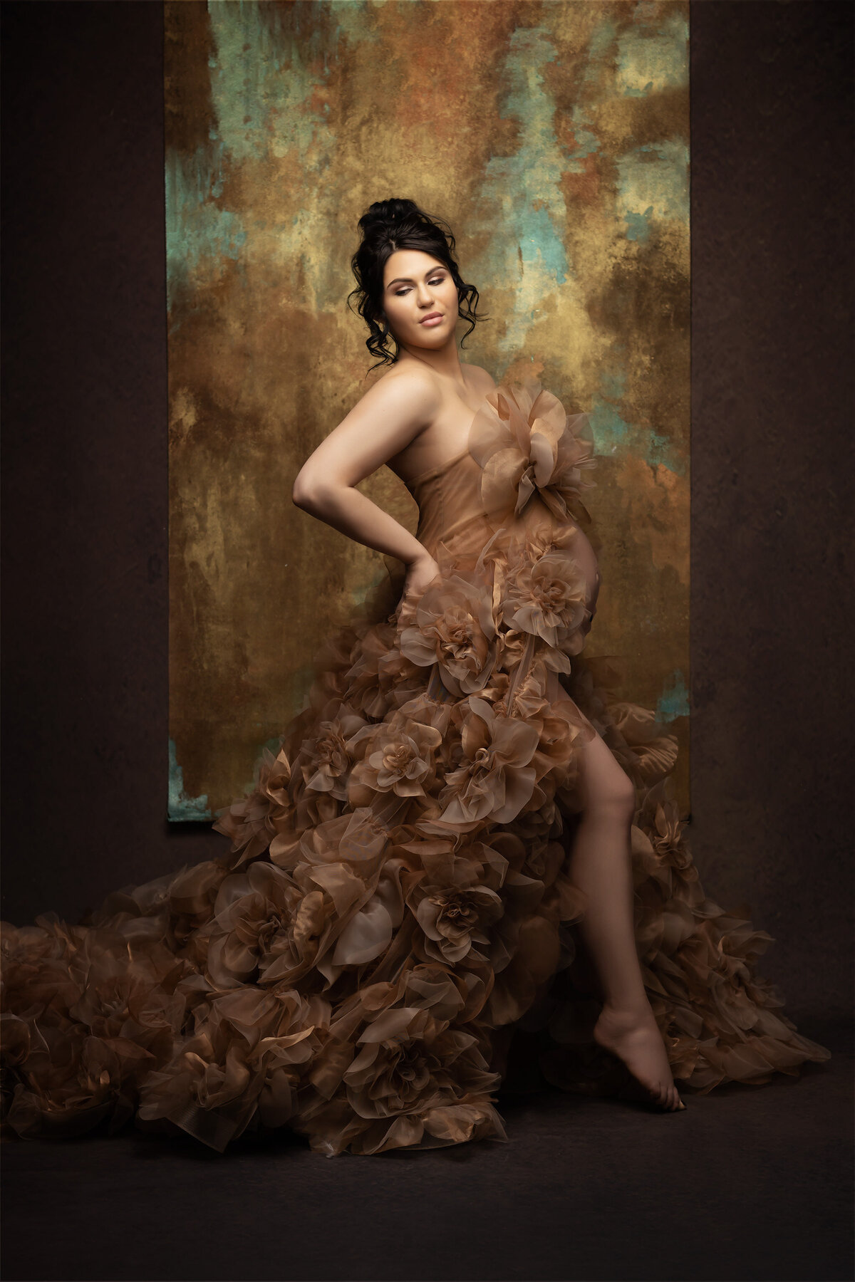 Pregnant woman in a luxurious floral gown posed elegantly against a textured gold and teal backdrop.