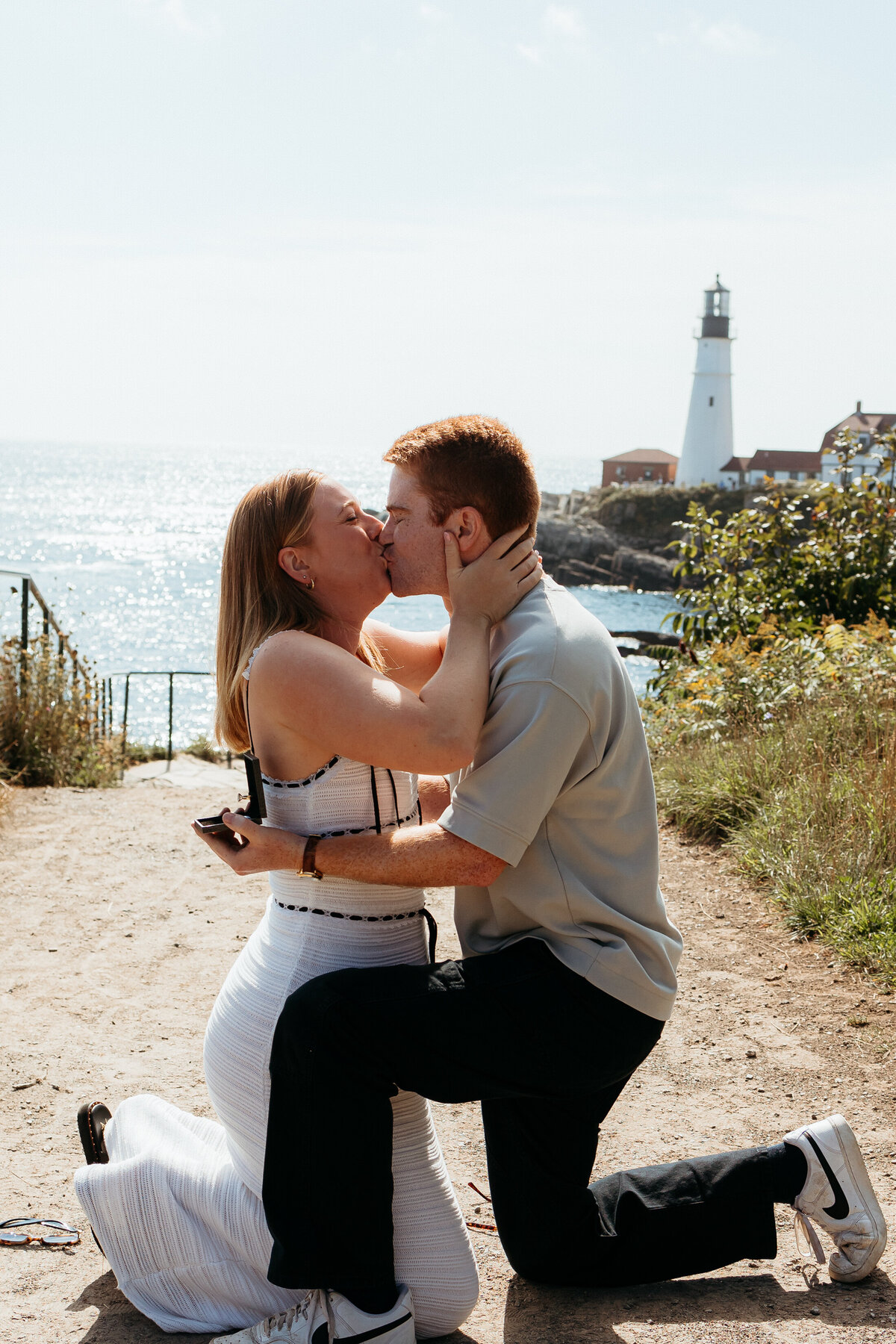 Portland Headlight proposal photos: couple kissing with the fiance holding the ring and lighthouse in the background.