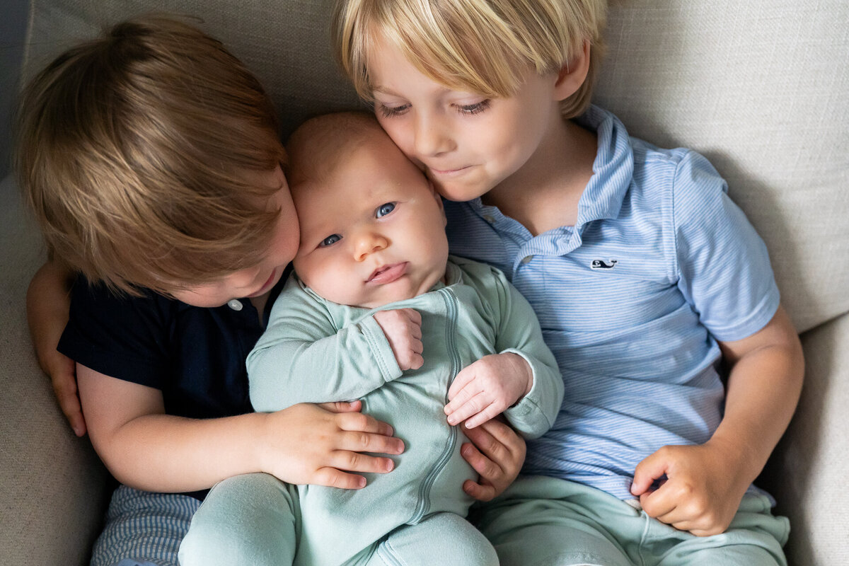 chubby newborn being held on  rocking chair by two older preschool brothers