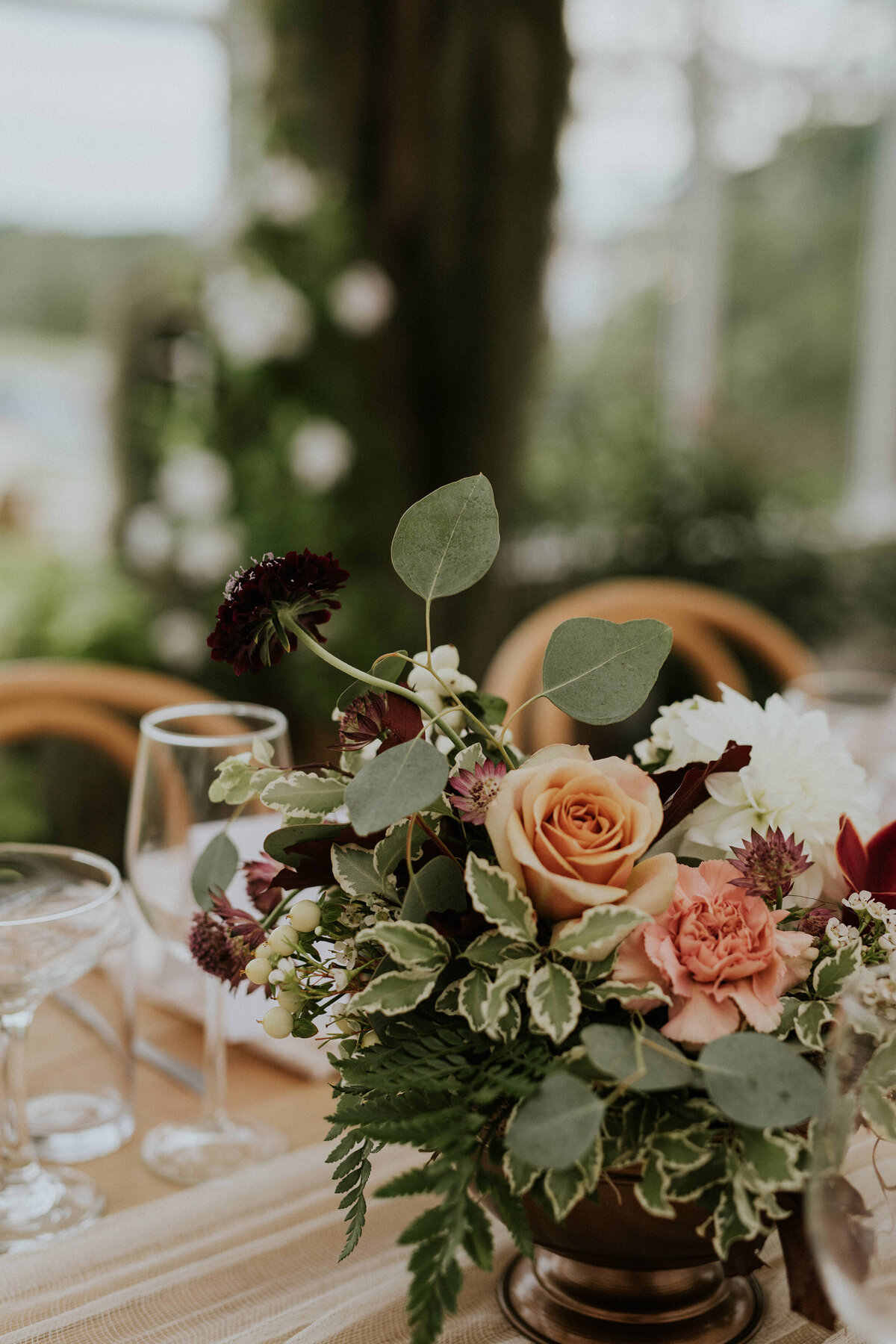 A wedding reception table with a centerpiece of white dahlias, dark purple scabiosa, mustard roses, peach carnations and mixed greenery in a bronze footed bowl