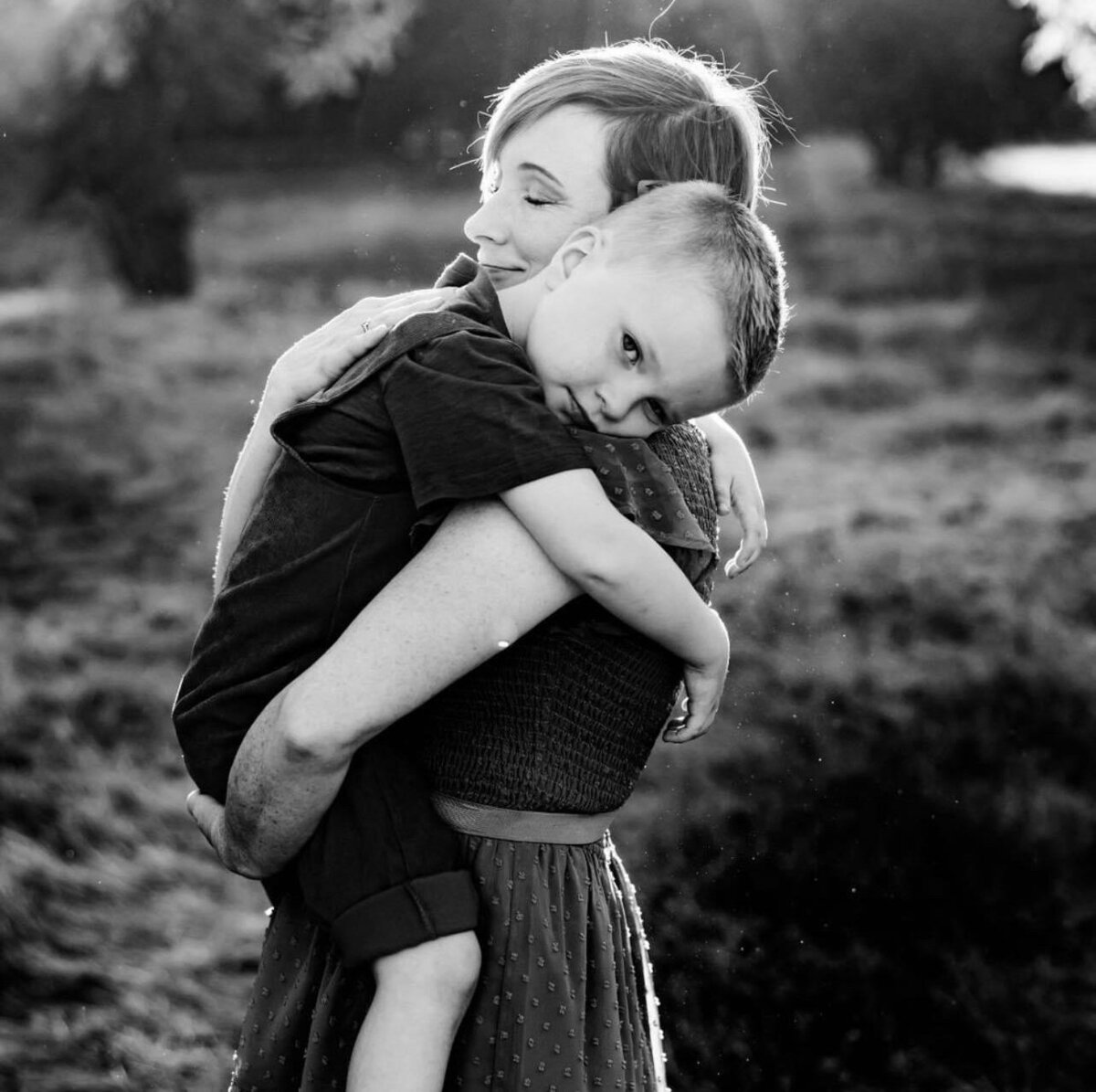 A mother holds her son, as he drapes his head over her shoulder and looks at the camera. She looks forward, eyes closed, smiling and embracing him.