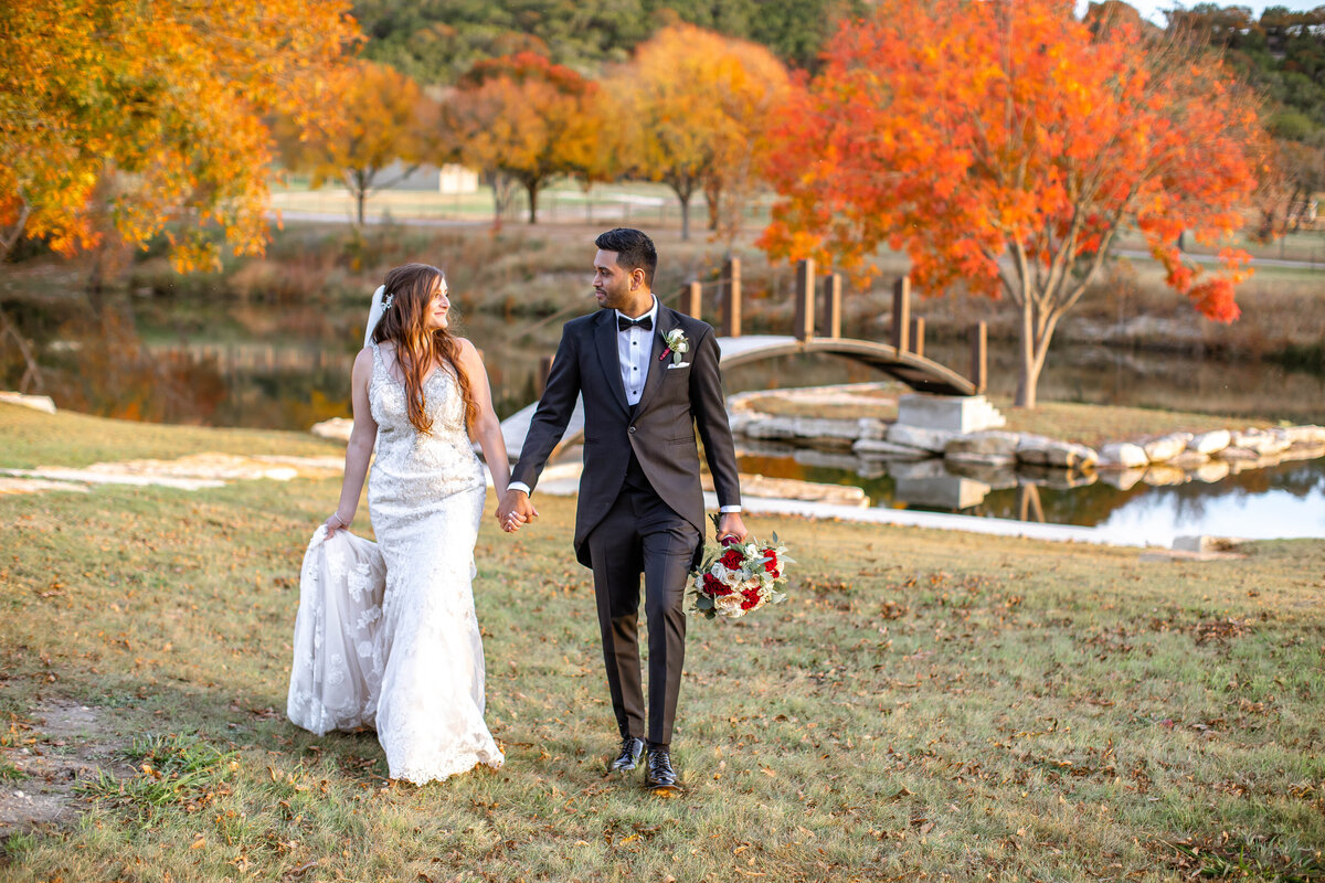 bride and groom gaze at each other while walking from lake bridge and Fall trees at Sendera Springs in Kerrville Texas