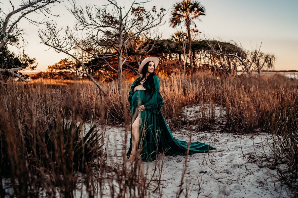 Pregnant mom cradling her belly and posing on sand at sunset in a grassy beach scene 