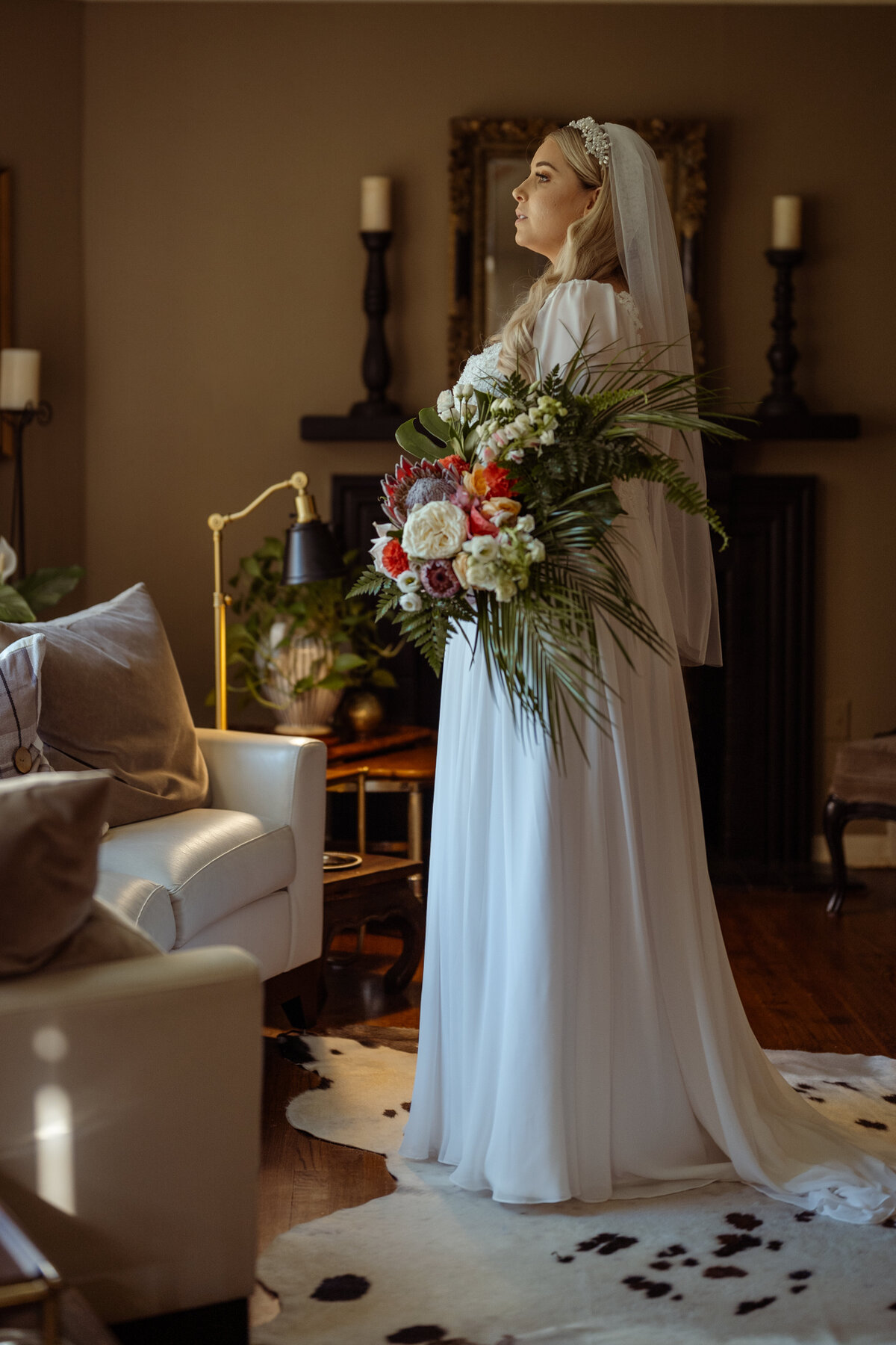Elegant bride in a vintage-style gown, holding a vibrant tropical floral arrangement, as she prepares for her wedding ceremony in Fort Worth. The bride stands gracefully in a warmly lit room, capturing the timeless beauty and unique style of her wedding day.