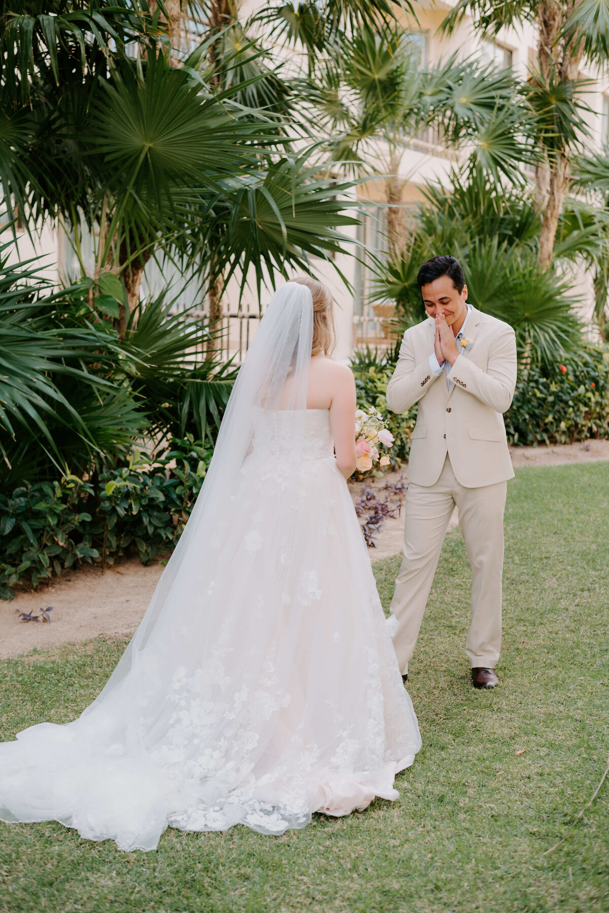 Groom reacts emotionally during a first look with his bride in a strapless lace wedding gown and veil, standing in a tropical garden setting.