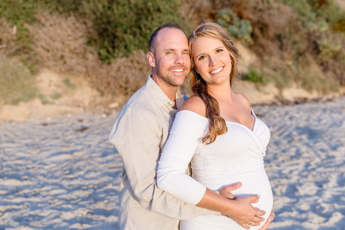 A couple stands on a sandy beach, smiling warmly at the camera. The man is embracing the woman from behind, and she is wearing a white dress, cradling her baby bump. The background shows sunlit bushes and sand.