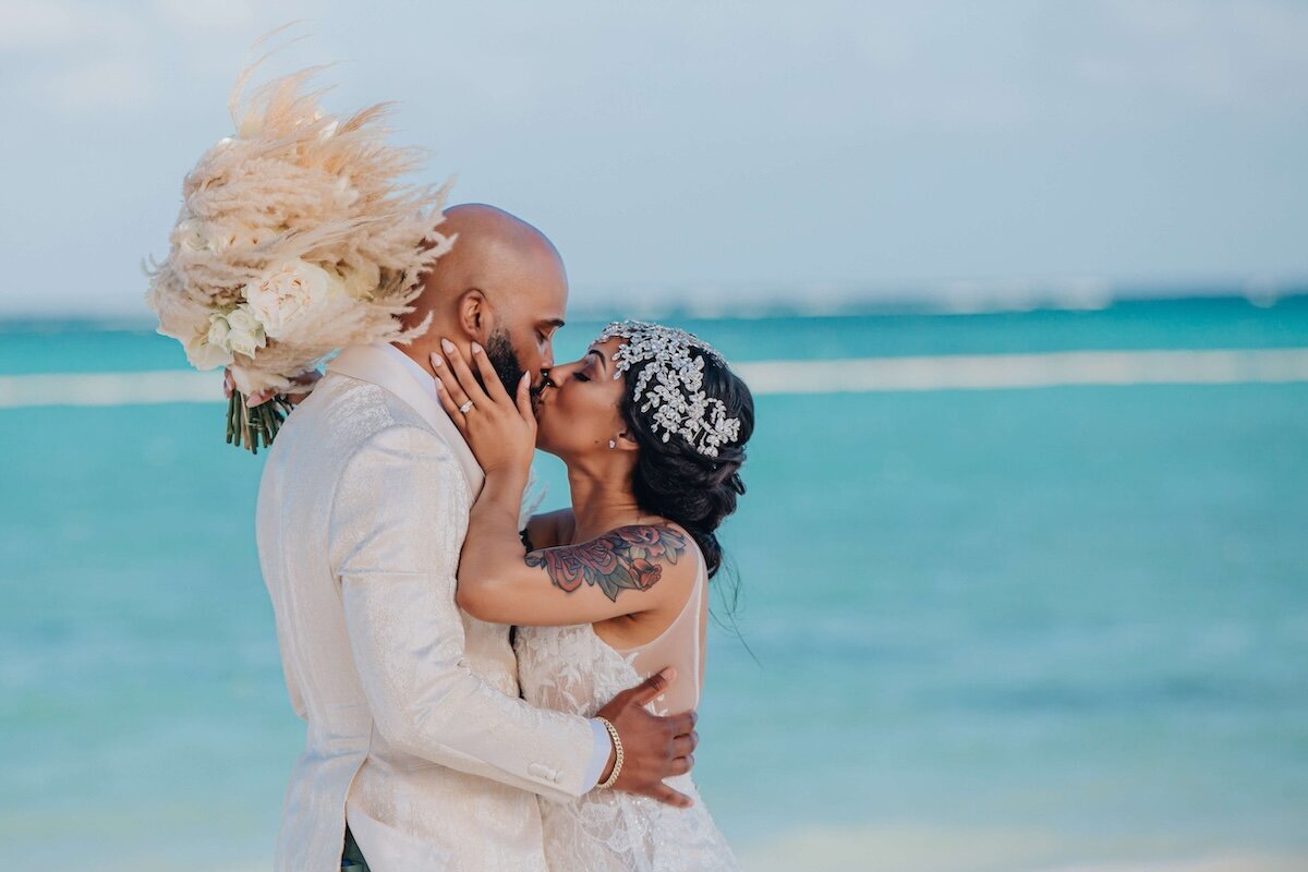 A bride and groom kiss on the beach, with turquoise ocean water in the background. The groom, dressed in a white suit, holds a bouquet of light-colored pampas grass, and the bride, wearing a jeweled headpiece, embraces him