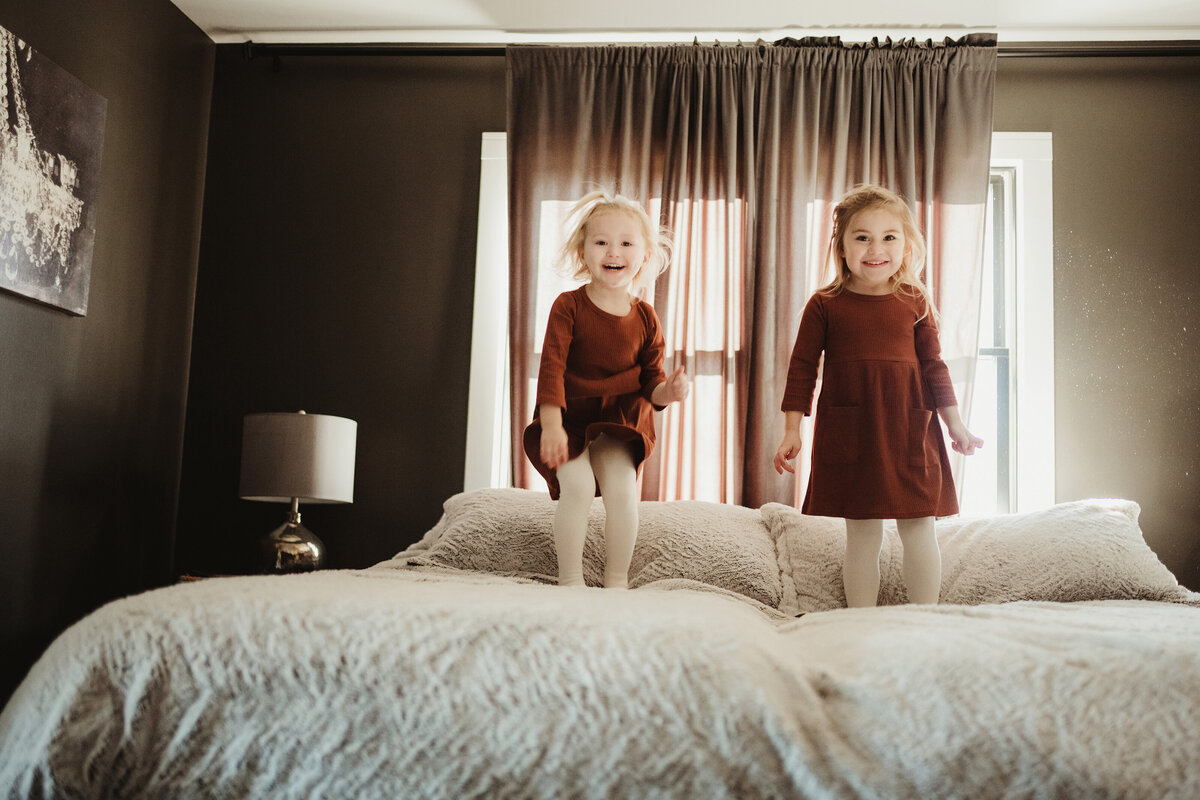 Two sisters are playing on the bed and looking at the camera.