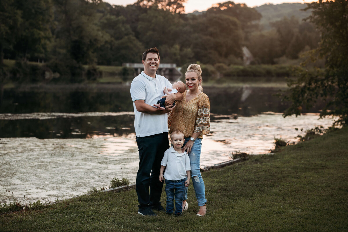 mom, dad, little boy and a baby standing in front of a pond standing in the grass at sunset
