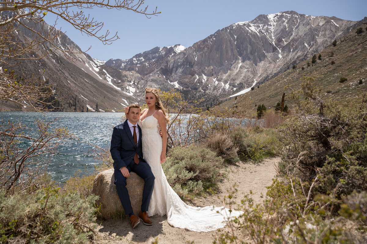 A groom sits on a rock as his bride leans against him on their California wedding day.