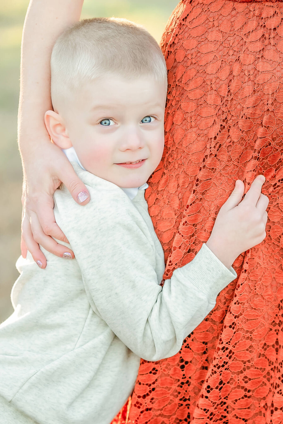 A young boy, wearing an off-white sweater, holds onto his mom's orange lace skirt during his Virginia Beach family photography session.