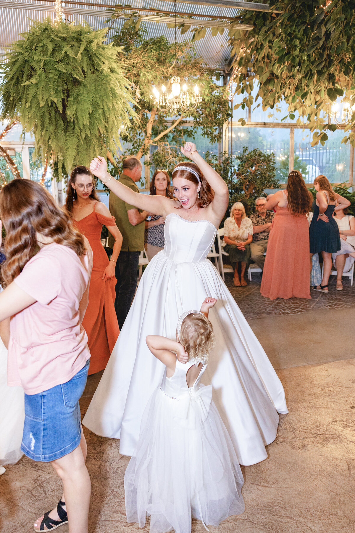 The bride throws her hands up in excitement while dancing with guests and a young flower girl at her wedding reception