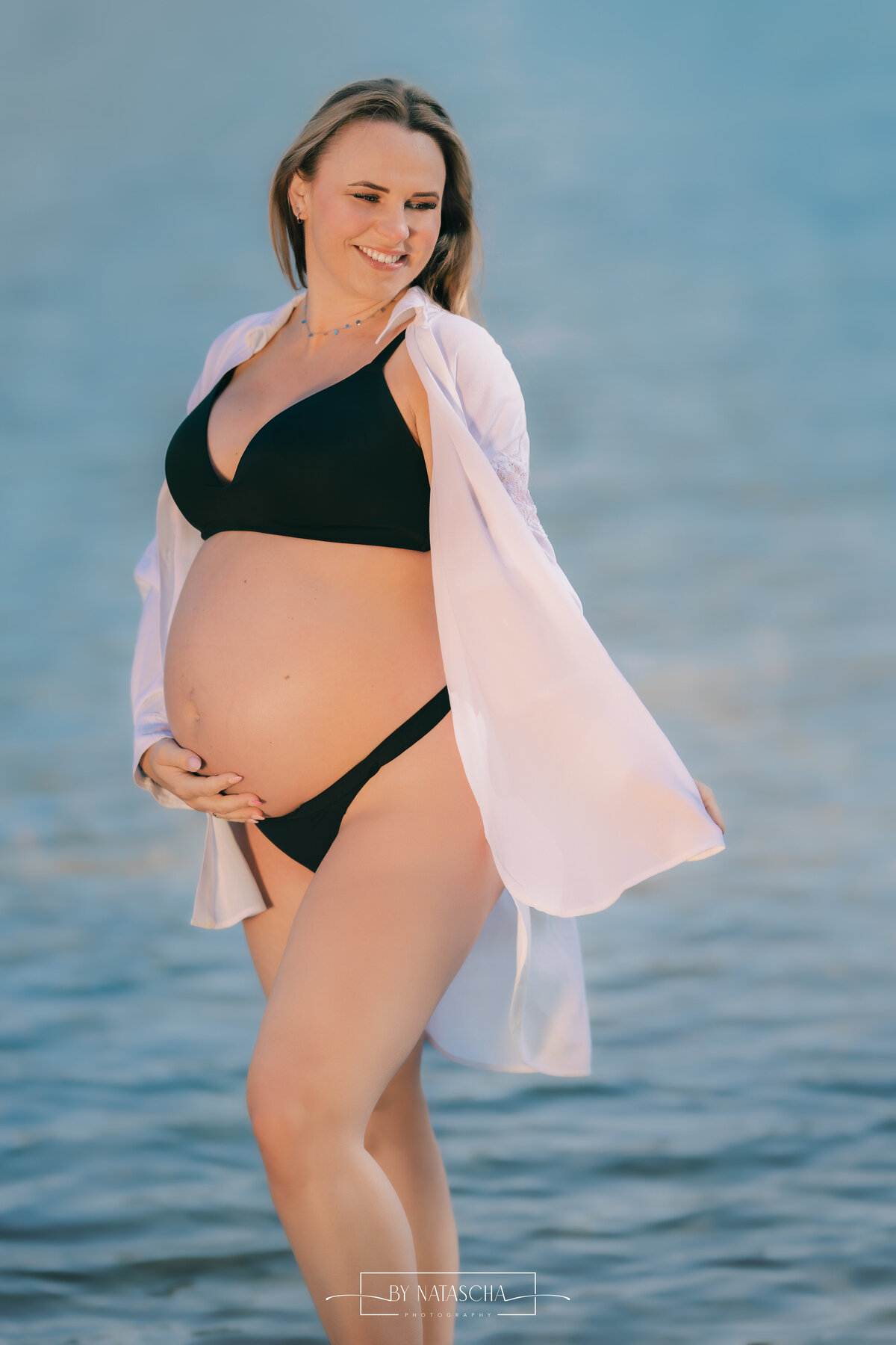 zwangere vrouw in lingerie met witte blouse met op de achtergrond helder blauw water en blouse waait open naar achteren