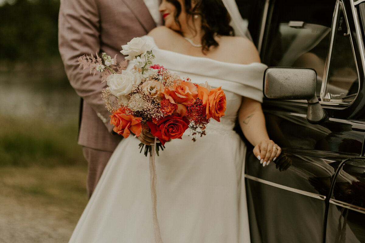 Mariés l'un contre l'autre, posés contre une voiture ancienne noire. Le marié porte un costume rose et la mariée son bouquet de fleurs rouges et oranges. Shooting photo réalisé en vendée par Laura Termeau.