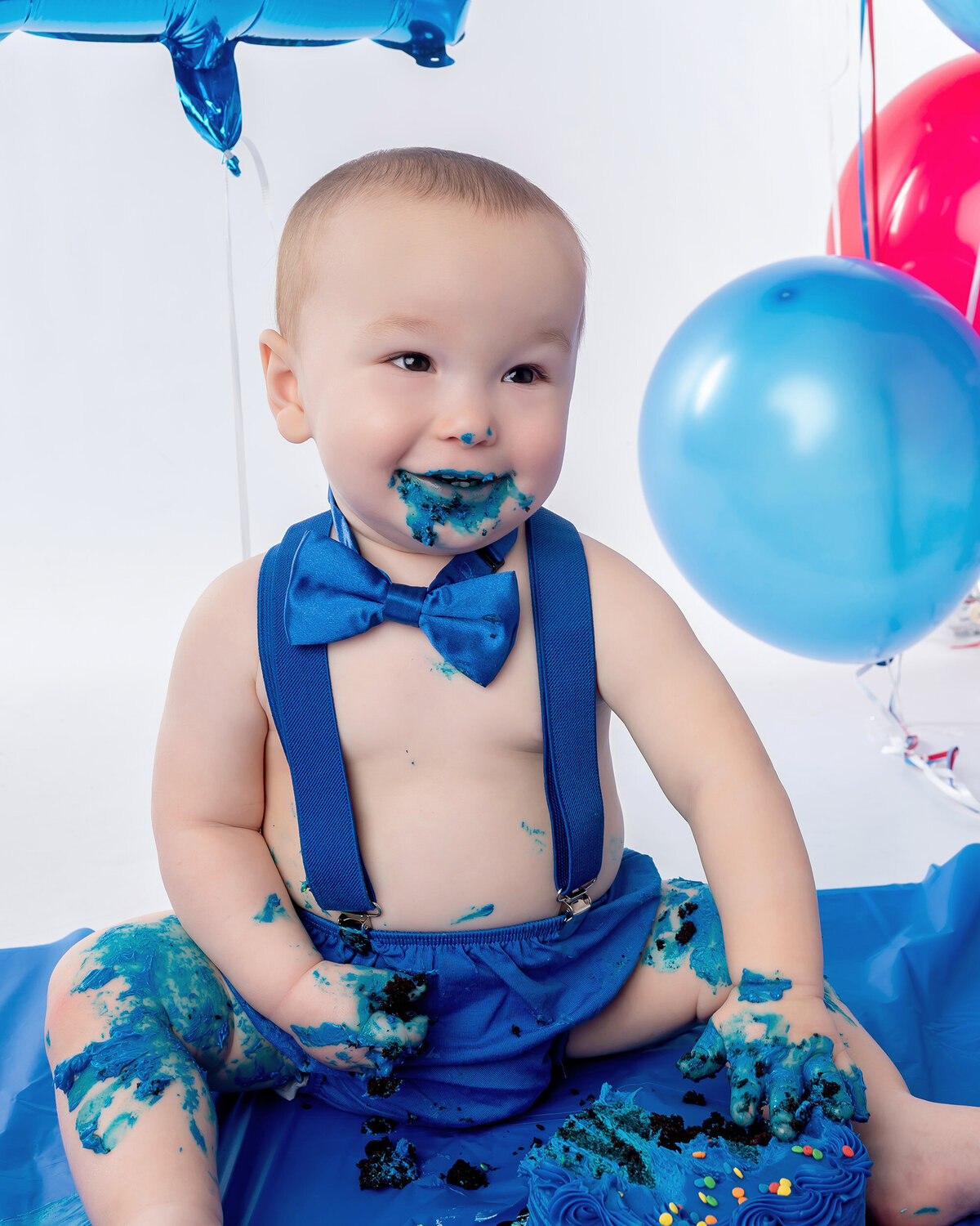 Baby in blue smiles as he sits with cake and balloons.