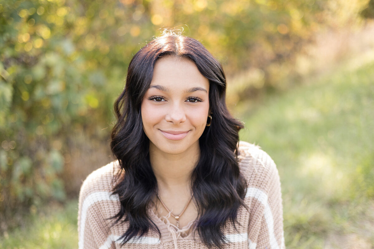 high school female senior, with long black hair, sitting, smiling softly at the camera wearing a light tan and white stripped shirt.