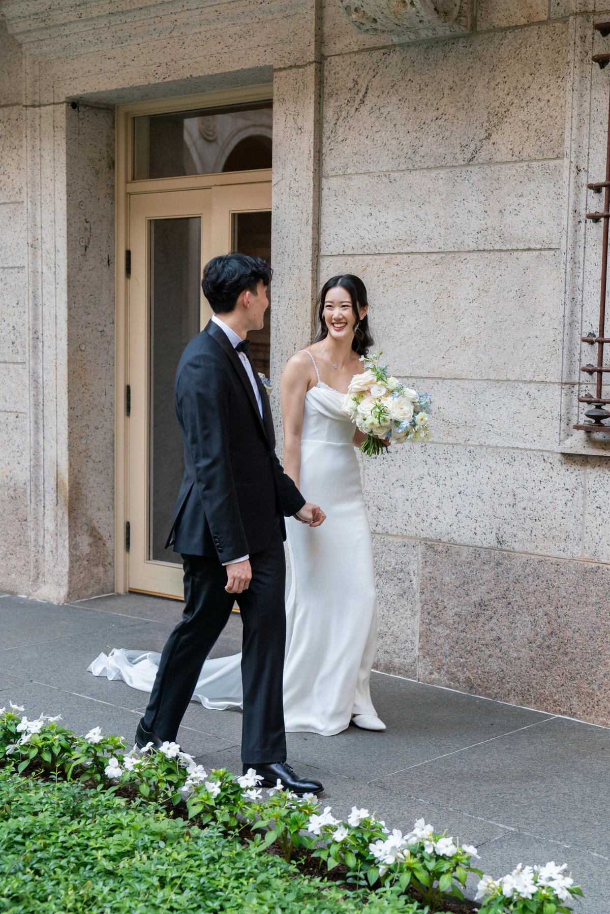 Rhode Island Wedding Photographers | Kelsey Sheehan Photography Timeless Rhode Island Weddings | A bride in a white dress smiles while holding a bouquet, walking with a groom in a black suit. They are outdoors against a stone wall background.