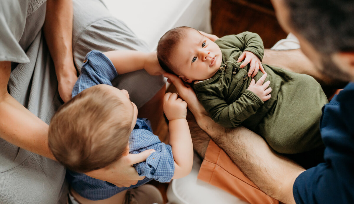 Newborn Photographer, dad holds baby in arms as older brother sits close to admire his sibling