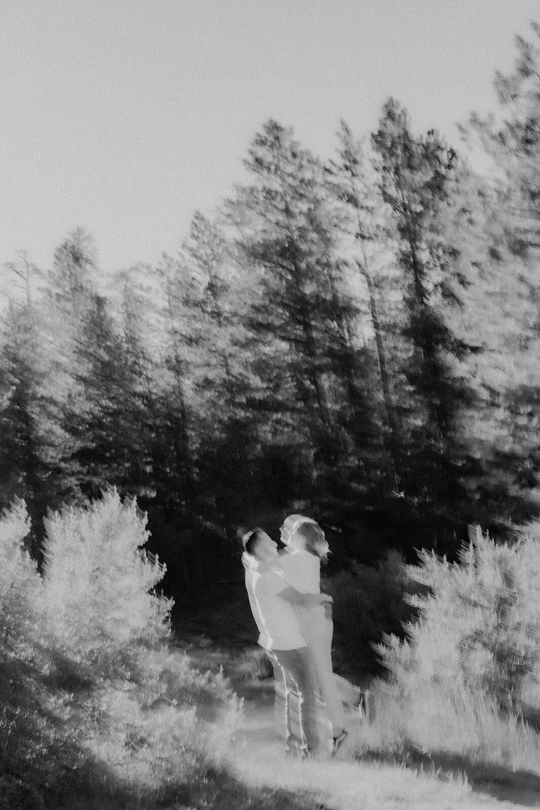 A black-and-white, motion-blurred image of the couple in a forest, appearing to embrace in a playful lift.