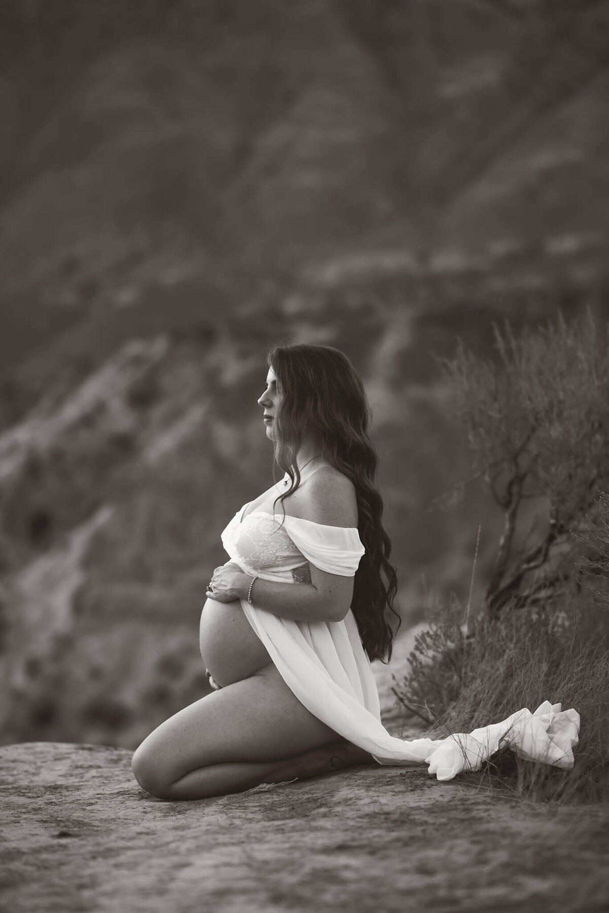 A black and white portrait of a pregnant mom kneeling at the edge of a cliff. She holds her belly and looks off into the distance.