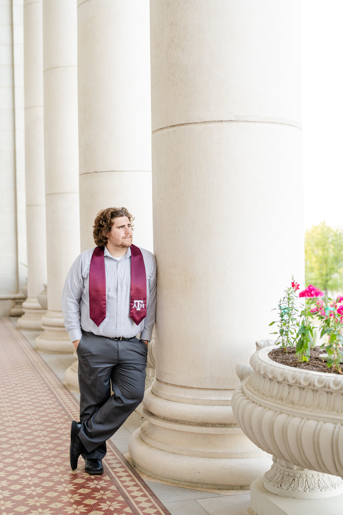 Texas A&M senior guy wearing maroon stole with button down and slacks while leaning against columns and looking out at Administration building