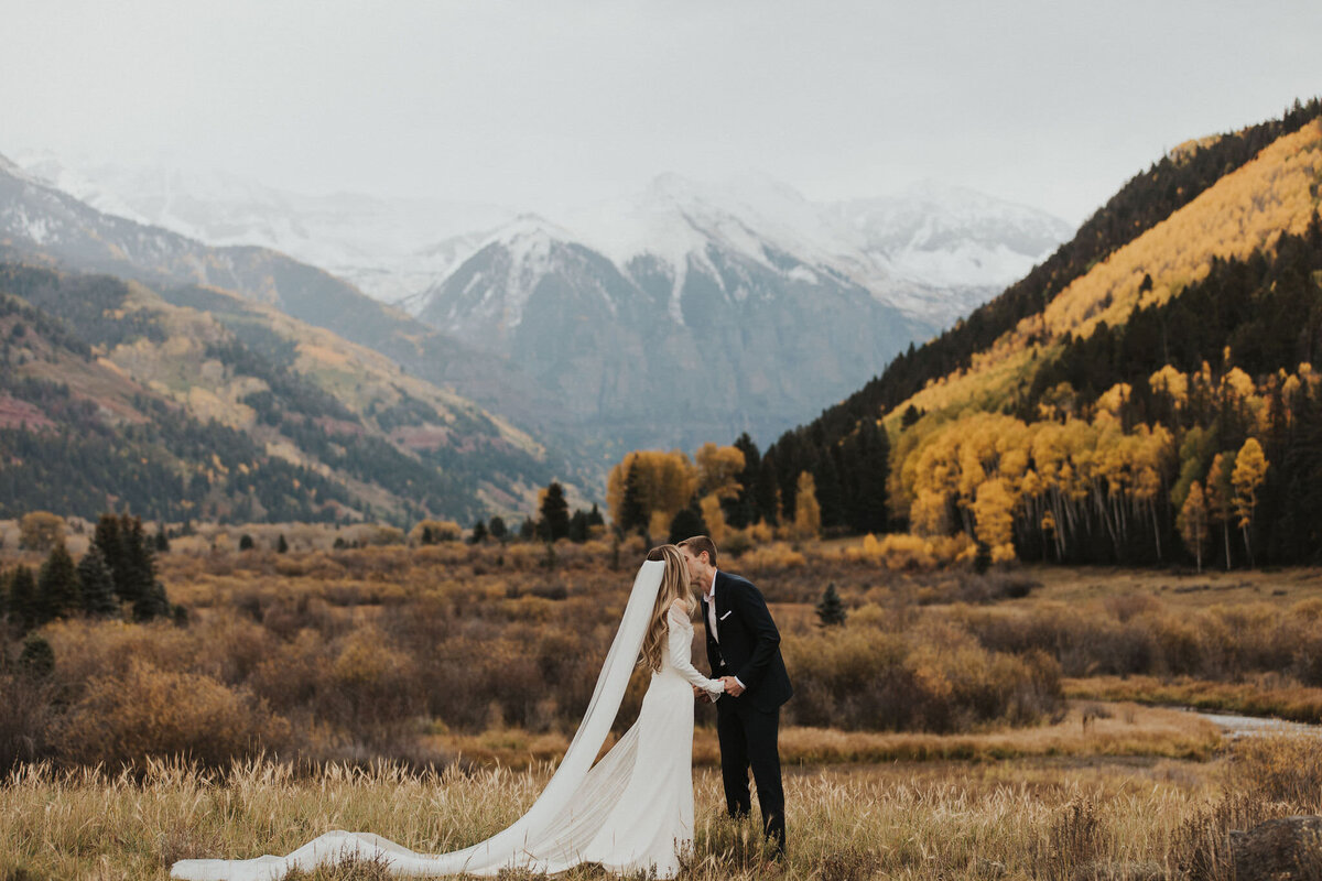 bride and groom standing in a field kissing with mountains in the background