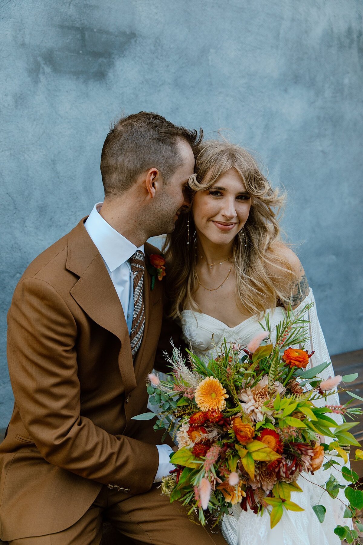 A boho bride in a strapless wedding dress with detached sleeves holds a large bouquet of orange and peach flowers with assorted greenery as the groom, wearing a brown suit kisses her cheek.