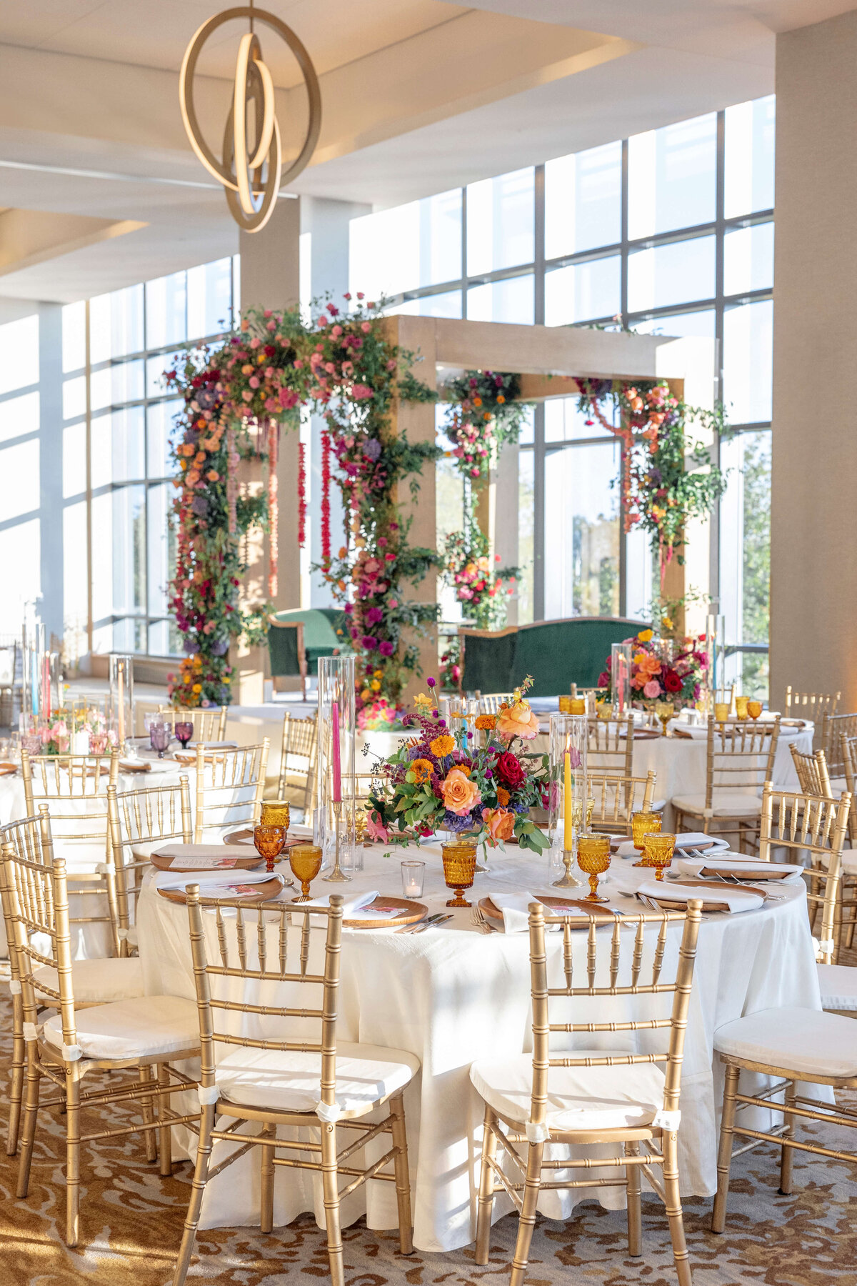 Elegant wedding reception with round tables adorned with white tablecloths, colorful floral centerpieces, and amber glassware. Gold Chiavari chairs surround the tables. A floral-accented wooden structure stands by tall windows in the background.