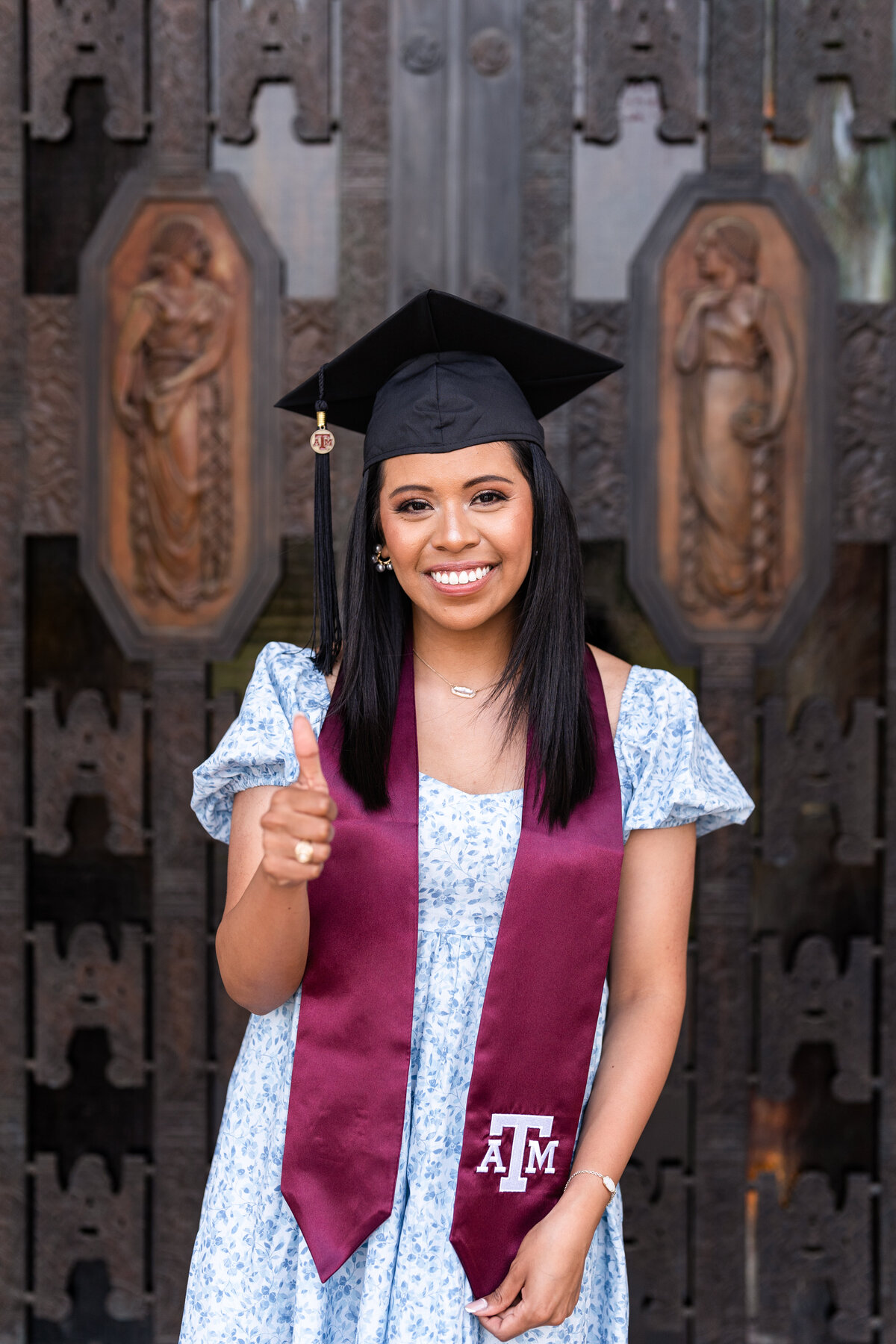 Texas A&M  senior girl holding stole and thumbs up wearing grad cap and blue dress in front of Administration building front door