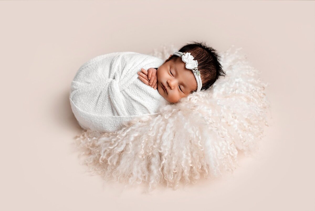 Baby girl in white wrap posed on a cream backdrop.
