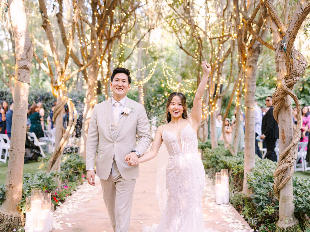 A joyful bride and groom walk down an outdoor aisle adorned with twinkling fairy lights and trees.