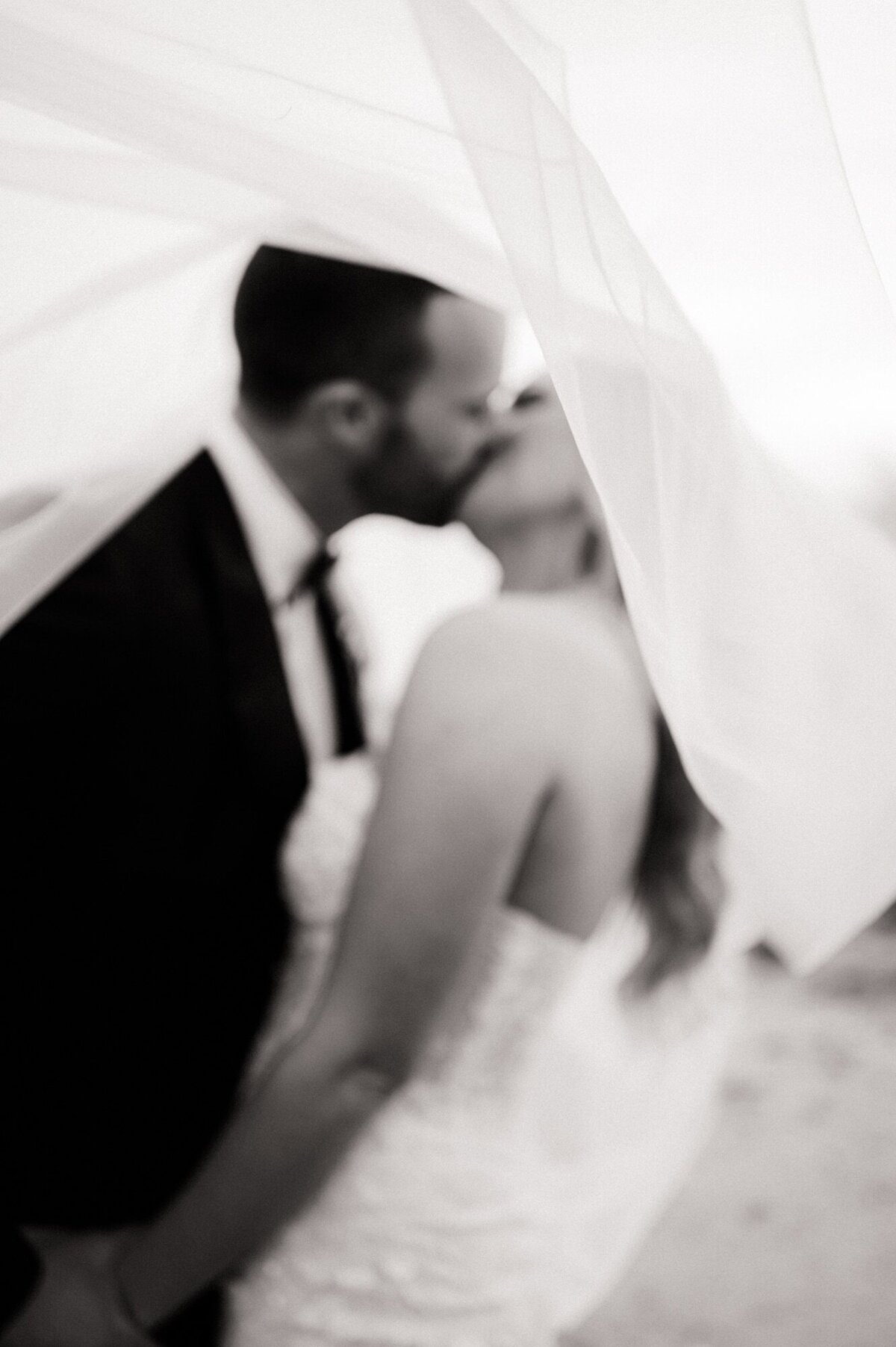 Bride and groom kissing under veil as wind blows veil