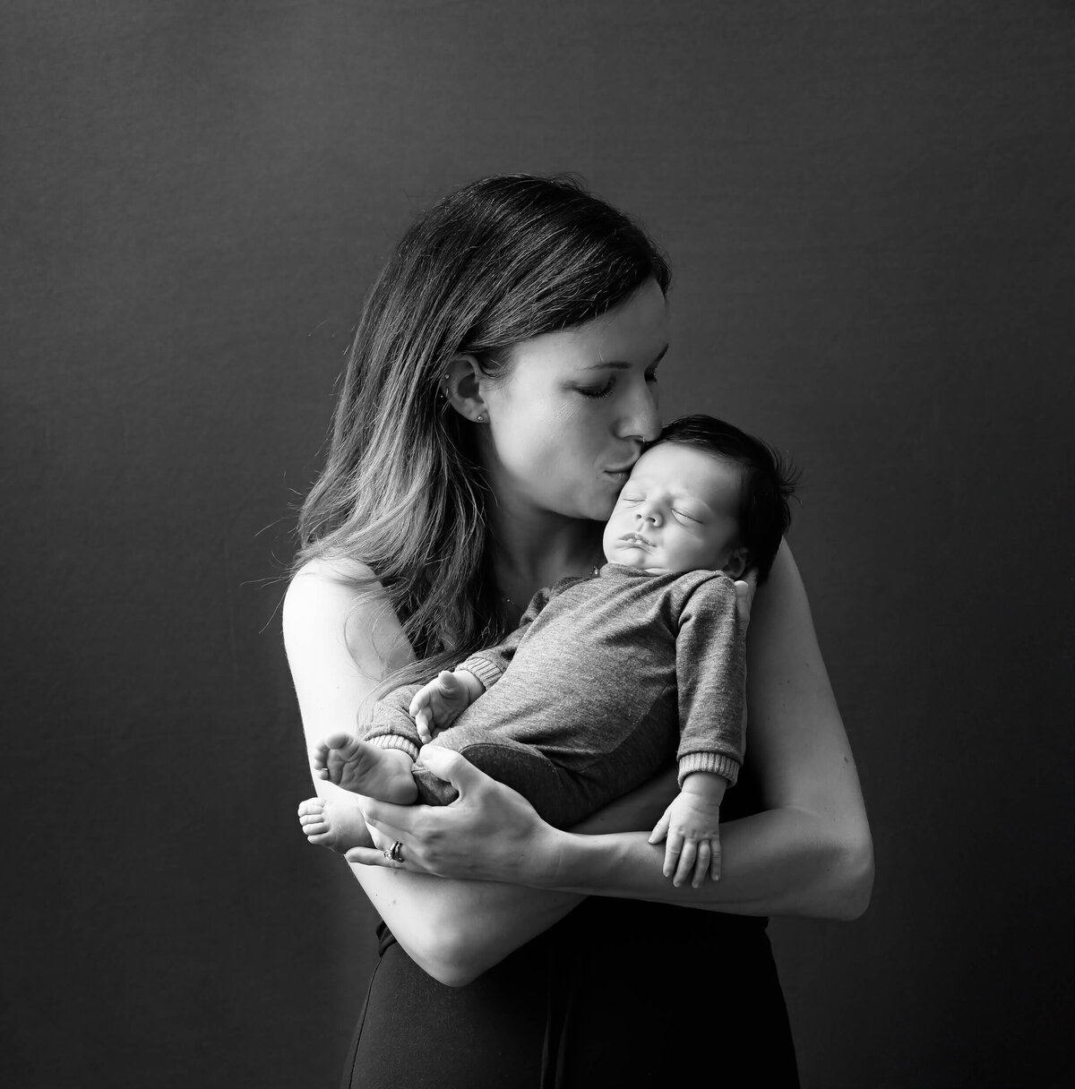 Black and white photo of new mom holding her newborn son and giving him a gentle kiss on his forehead. Photographed by E. Benecki Photography.