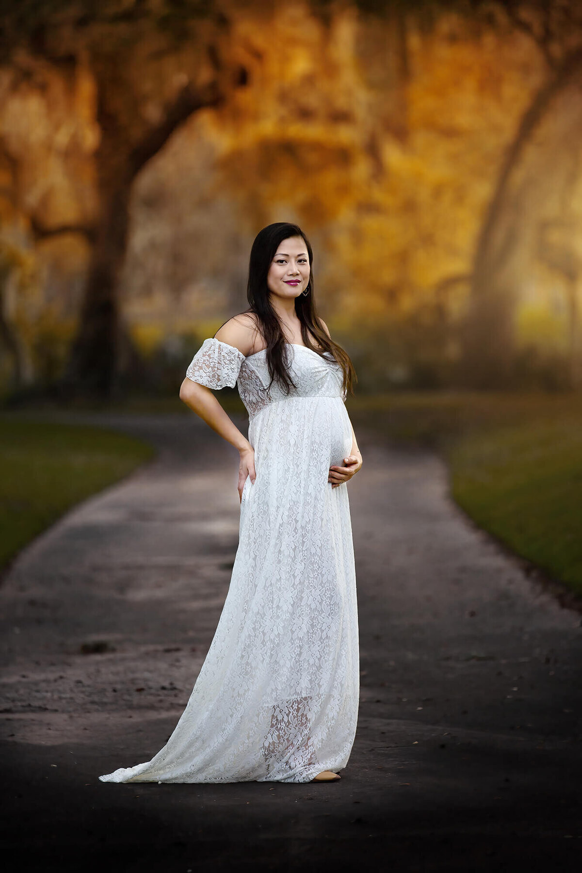 Beautiful pregnant lady with her hand on top of her belly wearing a lace dress with forest and trees in the background.