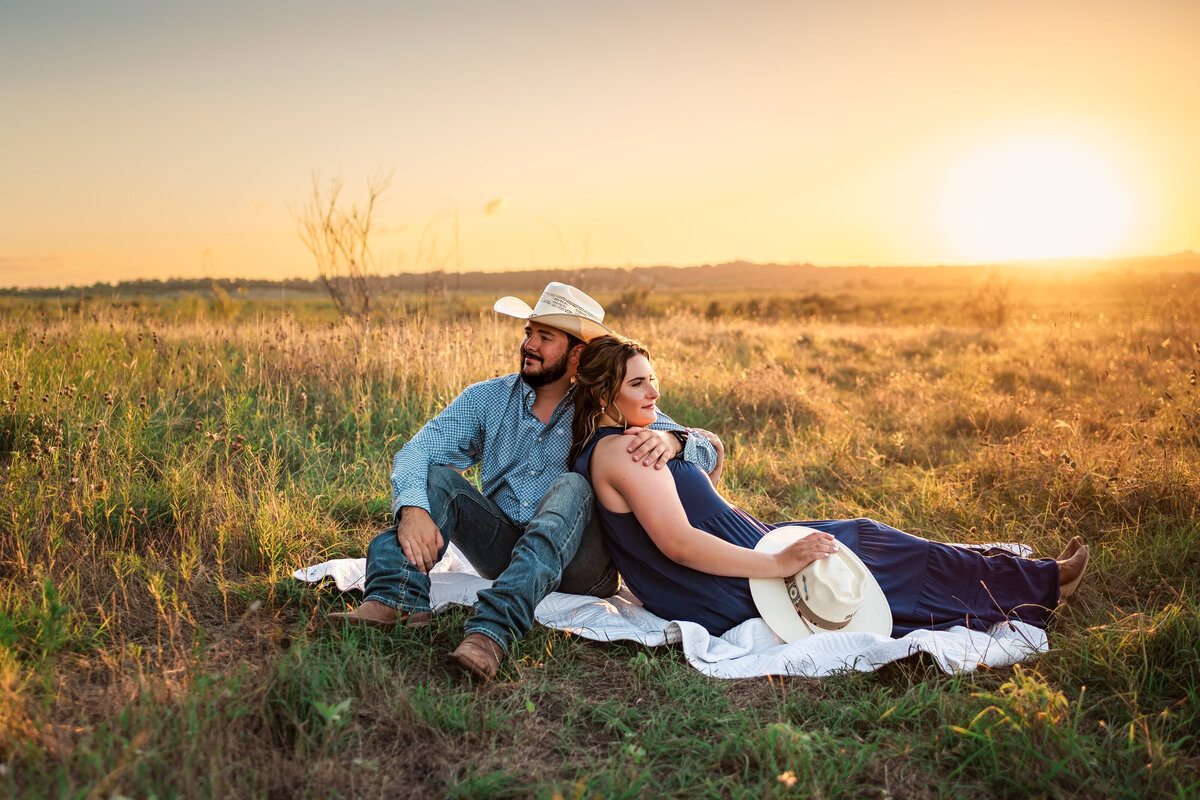 ngaged couple sitting on blanket in field at Harper Hills in New Braunfels, Texas, facing opposite directions during engagement session at sunset.