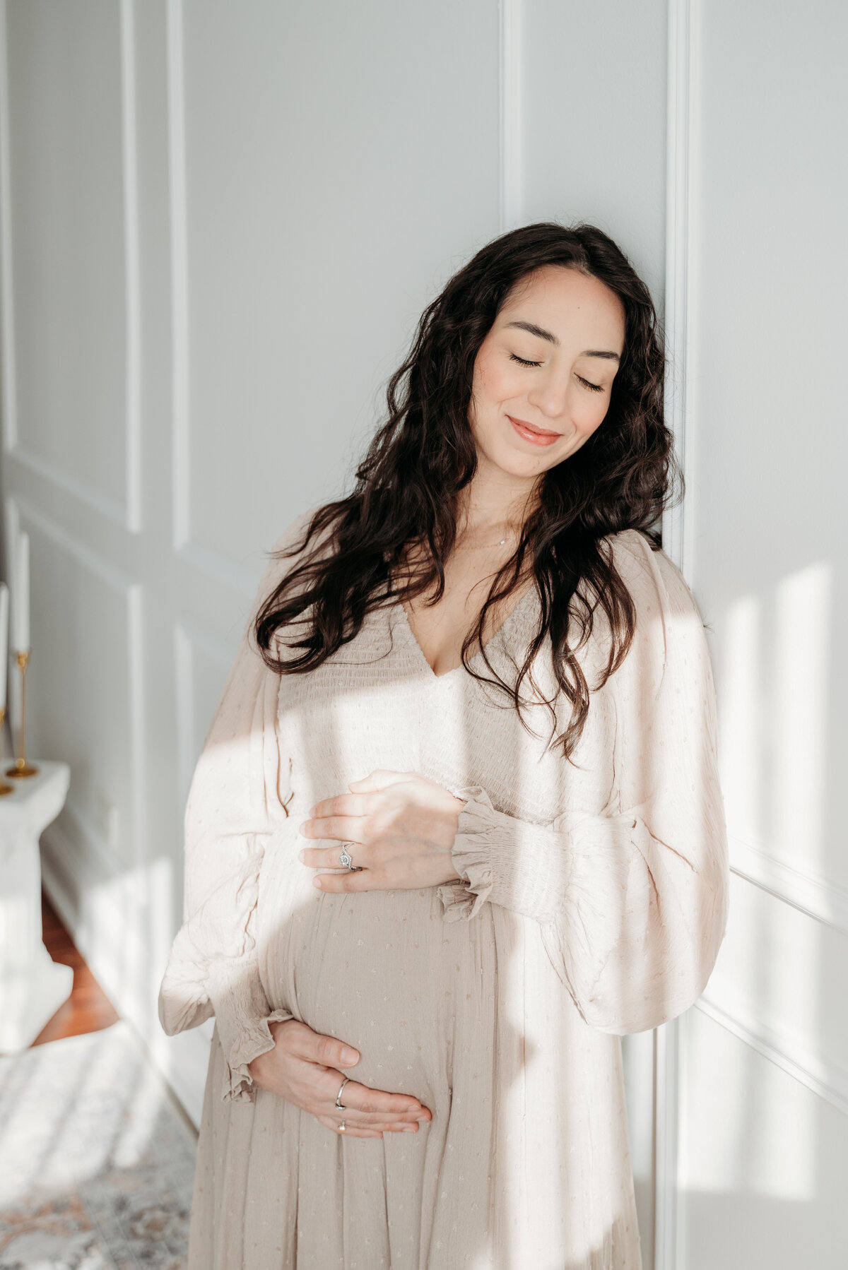 a glowing pregnant mother leans against the wall in a softly lit studio in Bartlett, IL. she turns towards the window, eyes closed, as golden light gently highlights her baby bump, a quiet, intimate moment of motherhood.
