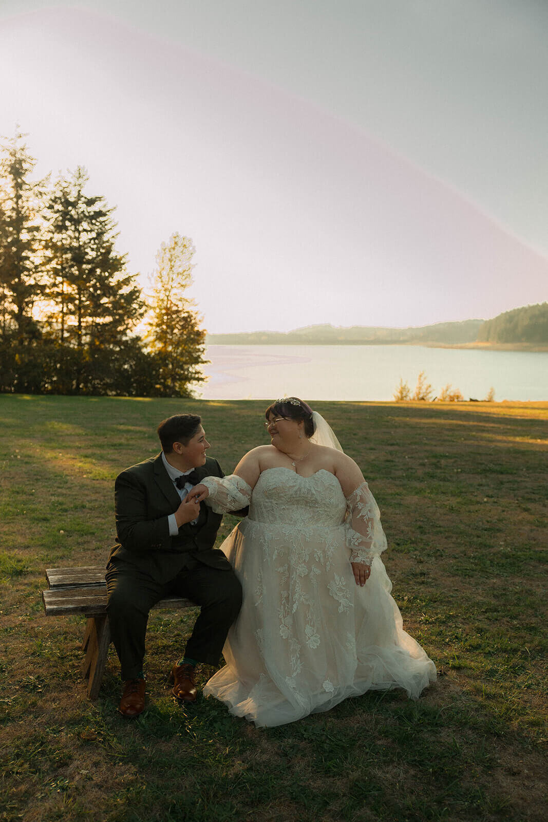 A wedding couple sits on a bench outdoors and smiles at each other with lake in background.