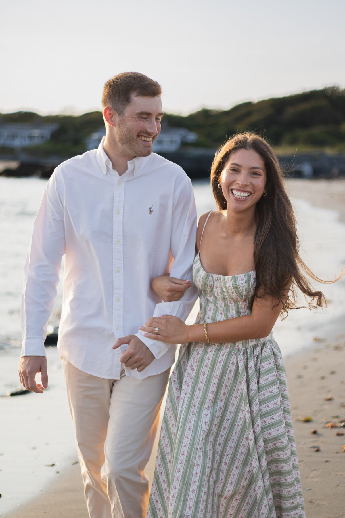 Collins Beach Newport RI | Kelsey Sheehan Photography Timeless Rhode Island Weddings | A smiling couple walks arm in arm on a beach at sunset. The man wears a white shirt and beige pants, and the woman is in a striped sundress.