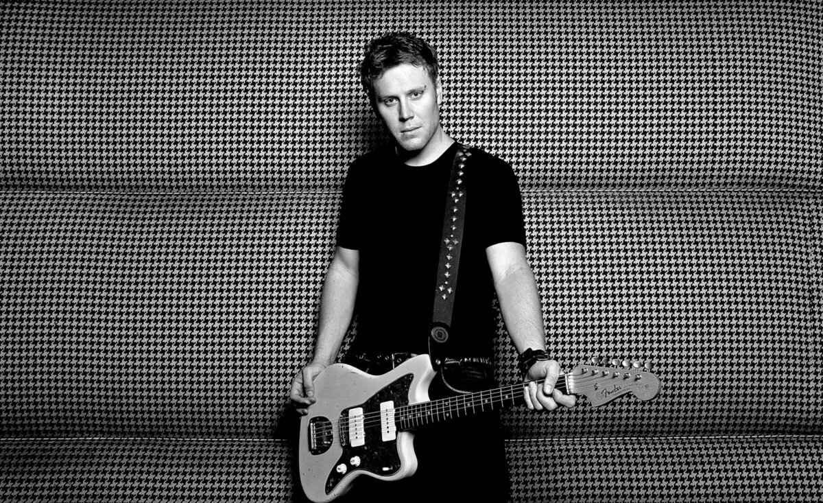 Male musician photo black and white Justin Wilke wearing black holding white electric guitar against patterned backdrop