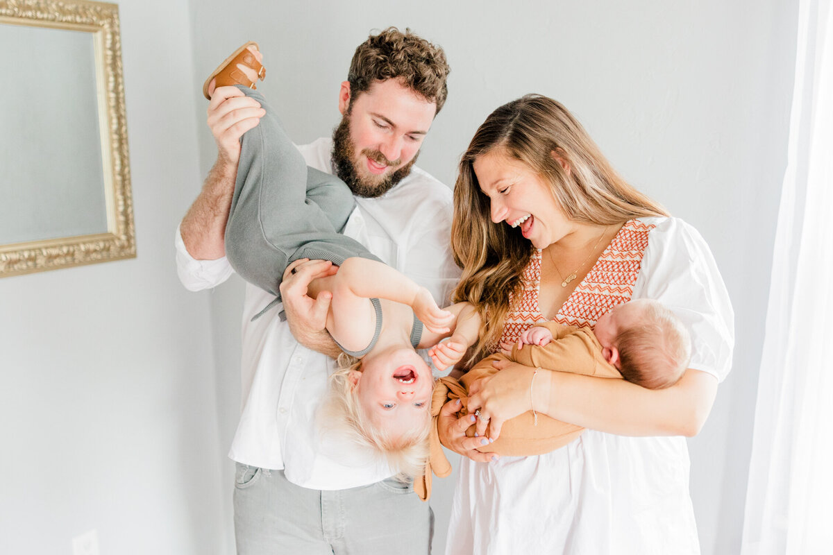 Family of four laughing with dad holding toddler upside down and mother holding newborn by Boston Newborn Photographer