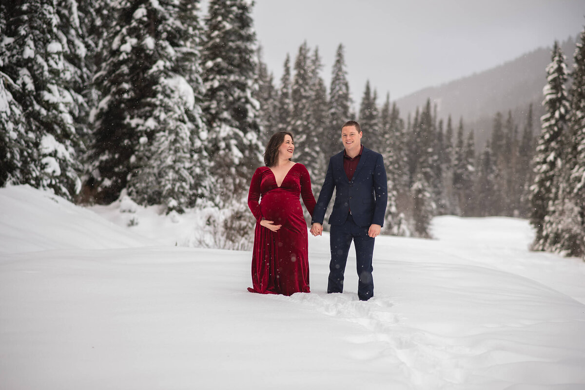 A happily married couple stand hand in hand in the snow. Husband is in a suit looking and smiling at the camera. Pregnant wife holds her belly and smiles at her husband. Portrait taken by Kamloops Photographer Brittany Danielle